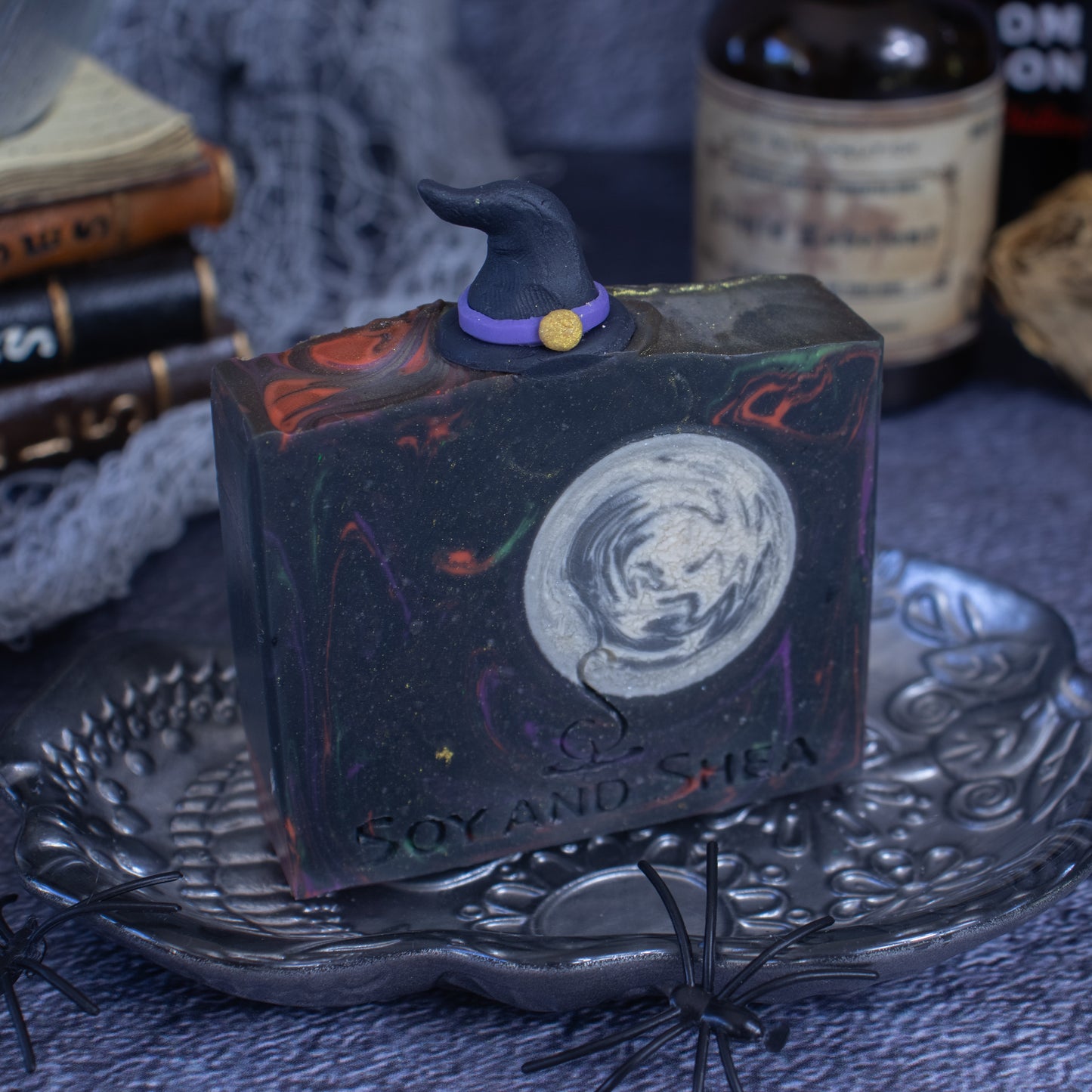 The Witching Hour Soap Bar (Halloween Edition) by Soy and Shea is a handmade block of soap decorated with a witch hat and moon design. Made with plant-based oils, it sits on a decorative tray surrounded by toy spiders. The presence of old books and a potion bottle in the background intensifies its mysterious scent and spooky atmosphere.