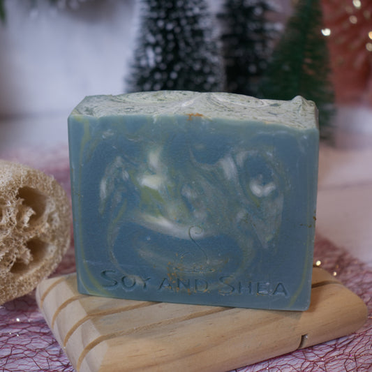 A Christmas Tree Soap Bar from Soy and Shea, featuring a marbled blue and white design, rests on a wooden soap dish. Decorative trees and a loofah sponge placed on pink netting create a festive ambiance in the background. This charming plant-based soap is enclosed in biodegradable wrapping, making it an ideal gift for those who are eco-conscious.