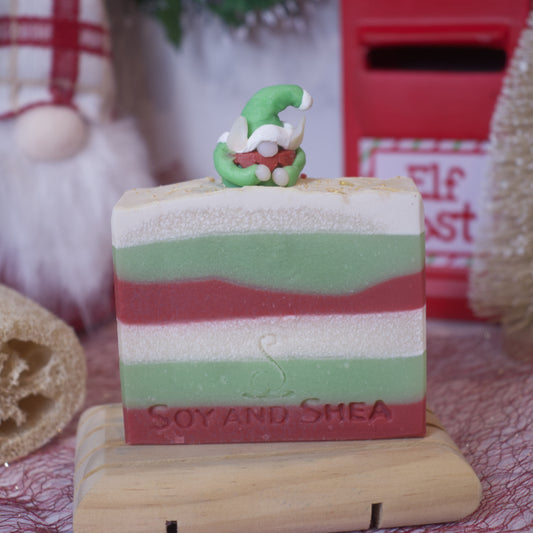 Displayed on a wooden stand is the Elf Magic Soap Bar (Christmas Edition) by Soy and Shea, featuring festive layers of green, white, and red. On top sits a small elf figurine. This handmade soap is crafted from plant-based oils and showcases an embossed logo, with holiday decorations in the background and a hint of Christmas aroma.