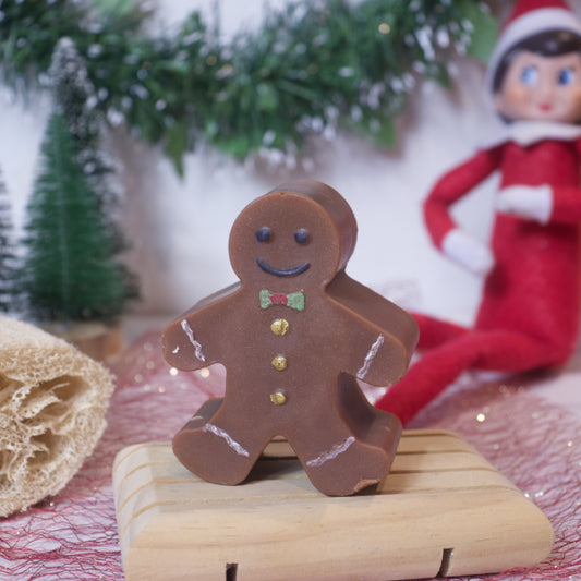 A cheerful holiday display features the Ginger Soap Bar (Christmas Edition) from Soy and Shea, designed as a gingerbread man with a charming smile, bow tie, and buttons. In the background, an elf doll dressed in red is nestled among green garland and tiny trees. A nearby loofah enhances the scene with its pleasant pairing of plant-based oils.