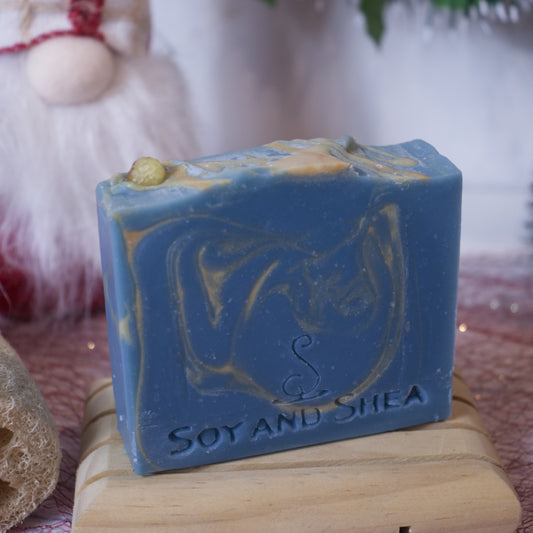 A blue and gold handcrafted Gold, Frankincense & Myrrh Soap Bar (Christmas Edition) by Soy and Shea, enriched with plant-based oils, is displayed on a wooden soap dish. The soap is embossed with the brand name "Soy and Shea." In the background, a soft-focus holiday decoration enhances the festive fragrance of the scene.