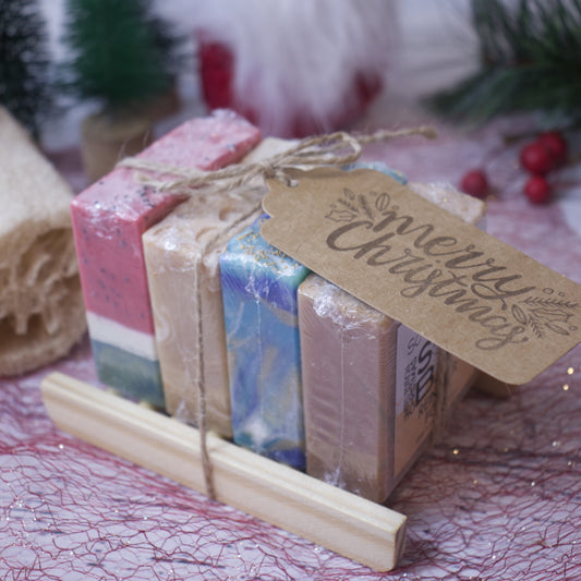 A Soap Bar Sampler from Soy and Shea's Christmas Edition, featuring vibrant handmade soaps tied with twine, sits elegantly on a bamboo dish. Accompanied by a rustic "Merry Christmas" tag, the set is adorned with festive touches such as berries and a small tree, making it an ideal gift for the holiday season.