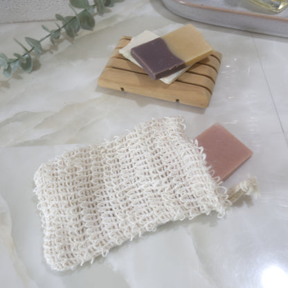 A cream, loosely woven cotton bag lies on a marble surface with a small piece of pink soap coming out the top.  To the back of the counter is a wooden soap dish with two small pieces of soap