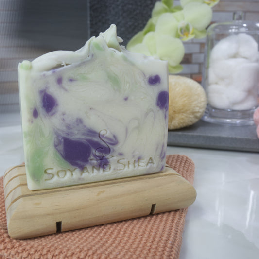 A white bar of soap with purple and green swirls on a wooden soap dish on a bathroom counter top