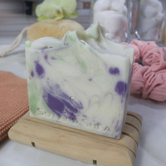 A white bar of soap with purple and green swirls on a wooden stand, surrounded by bathroom accessories