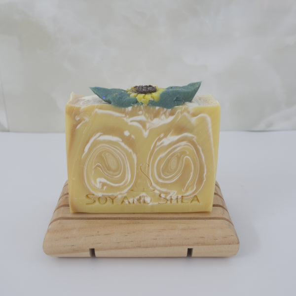 A yellow Bar of soap with a decorative sunflower on top, placed on a wooden stand against a white background.  The soap has a white and gold swirl through it
