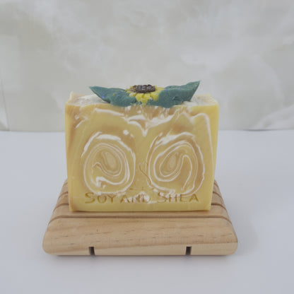 A yellow Bar of soap with a decorative sunflower on top, placed on a wooden stand against a white background.  The soap has a white and gold swirl through it
