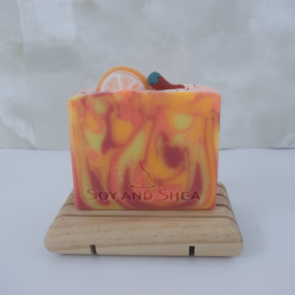 A Sweet Orange Chilli Pepper Soap Bar by Soy and Shea featuring yellow, orange, and red swirls sit on a wooden soap dish. The soap is decorated with an round orange slice and chilli made with soap