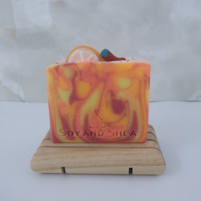 A Sweet Orange Chilli Pepper Soap Bar by Soy and Shea featuring yellow, orange, and red swirls sit on a wooden soap dish. The soap is decorated with an round orange slice and chilli made with soap