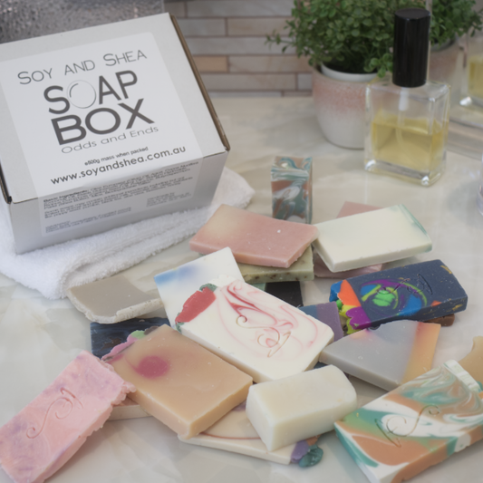 A white box with a label reading SOAP BOX ODDS AND ENDS sits behind a pile of soap pieces in various shapes and colours on a bathroom counter top with perfume bottle and potted plant.