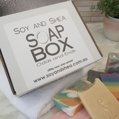 A white box with a label reading SOAP BOX ODDS AND ENDS sits on a white towel along with pieces of soaps in various colours and shapes