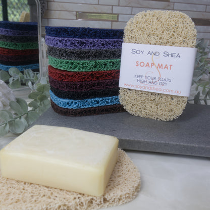A colourful stack of soap mats on a grey stone tray with a beige mat standing upright next to them with a product label.  To the front is another beige mat on the marble countertop with a cream soap resting on top.  The stack of soap mats are reflected in a hand mirror which rests beside the stack