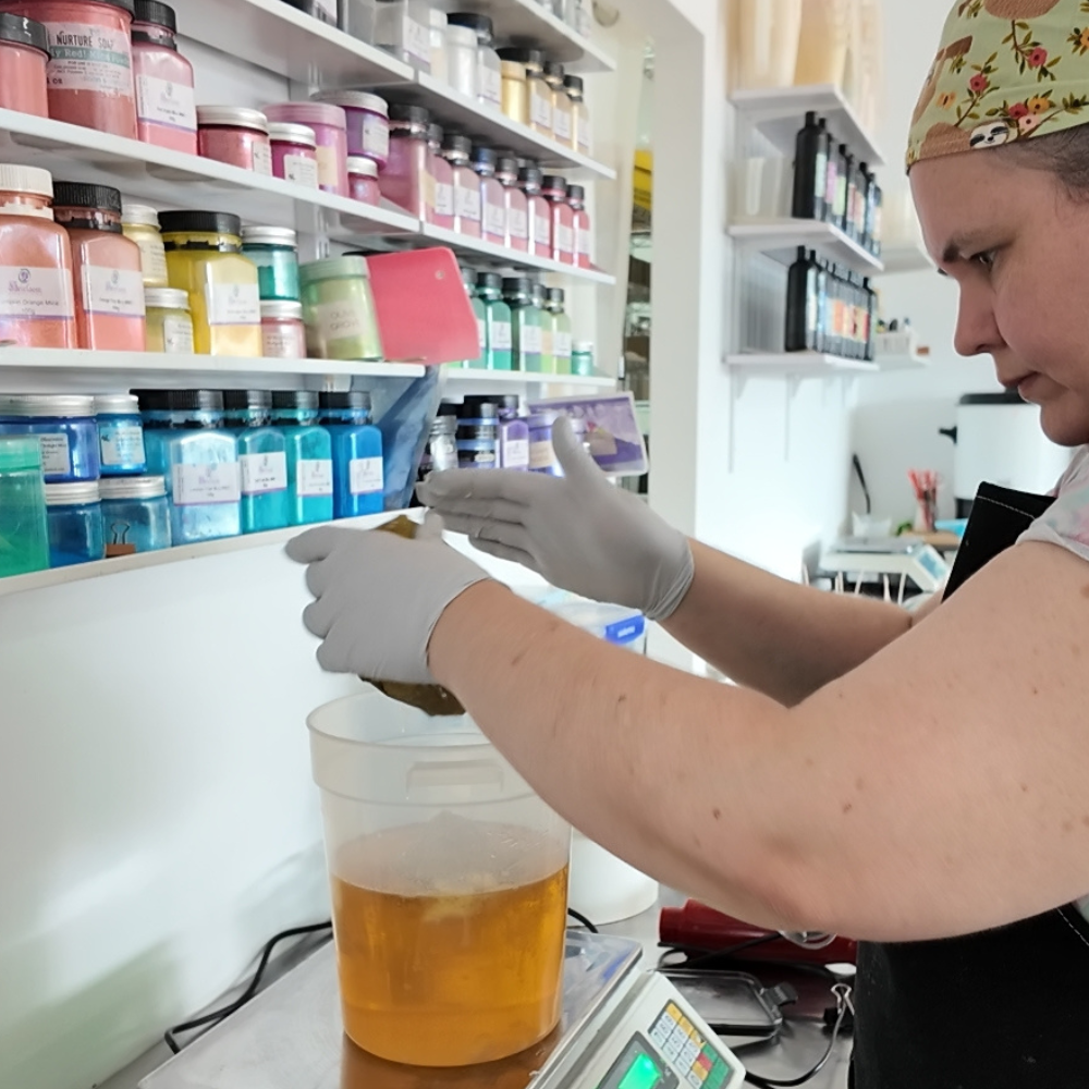 Keeley, the owner of Soy and Shea, is weighing something into a bucket filled with oils.  She is surrounded by bright posts of colour