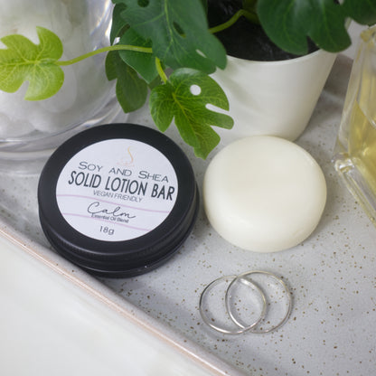 A small black round tin sits on a stone tray with a label reading SOLID LOTION BAR.  Next to it is a solid white bar of lotion.  The items are surrounded by jewellery, a potted pland and a bottle 