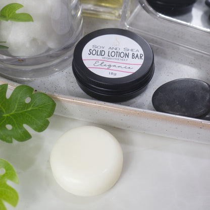 A small round tin with a label reading SOLID LOTION BAR sits on a stone tray surrounded by greenery and a jar of cotton balls.  In front of the tray is a solid round white lotion bar.