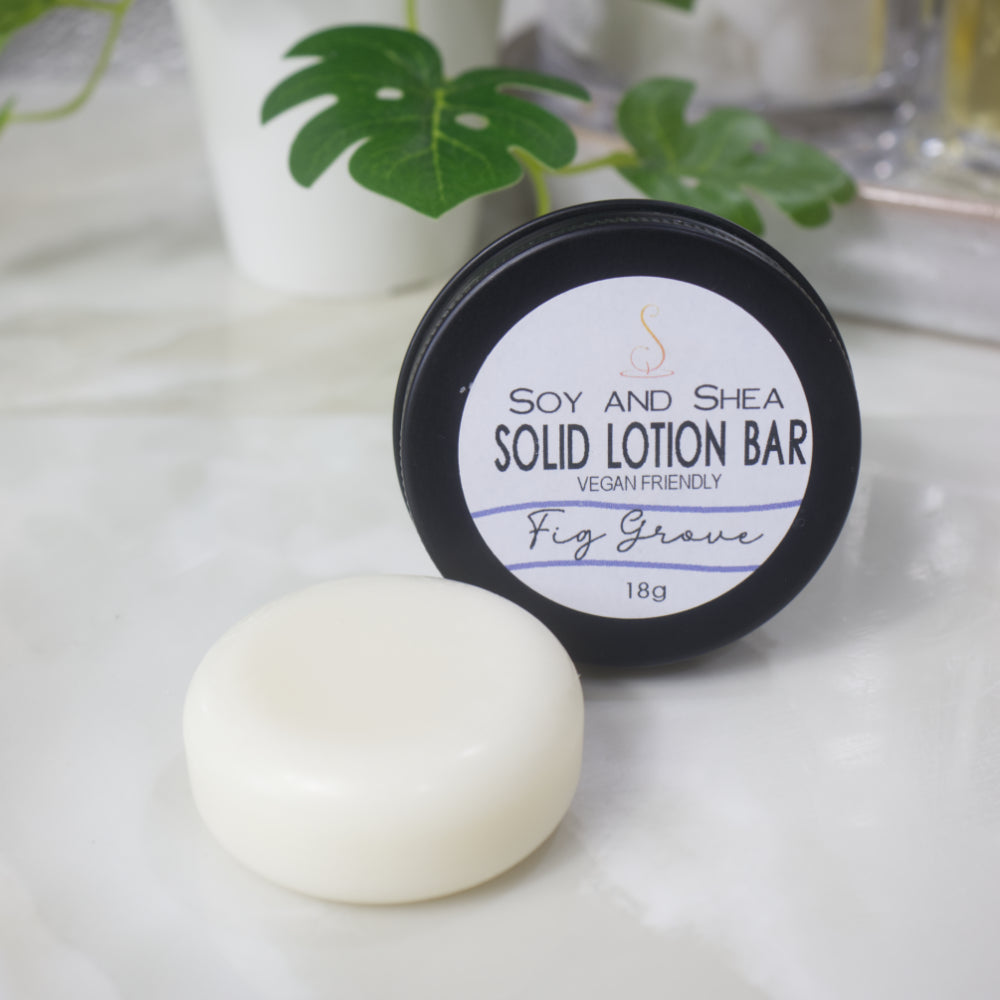 A small black tin with a label reading SOLID LOTION BAR stands on its side with the label facing forward.  In front of the tin is a solid white, cylindrical puck of lotion.  The background is blurred with greenery.