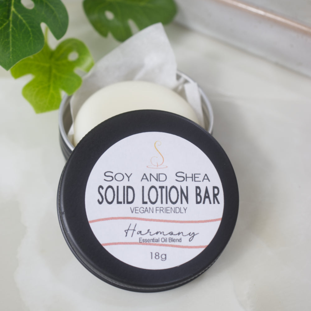 A small black round tin sits on a marble surface with a greenery to the back left of the overhead shot.  The lid of the tn has been removed and rests on the side revealing a solid white lotion nestled in some paper inside the tin