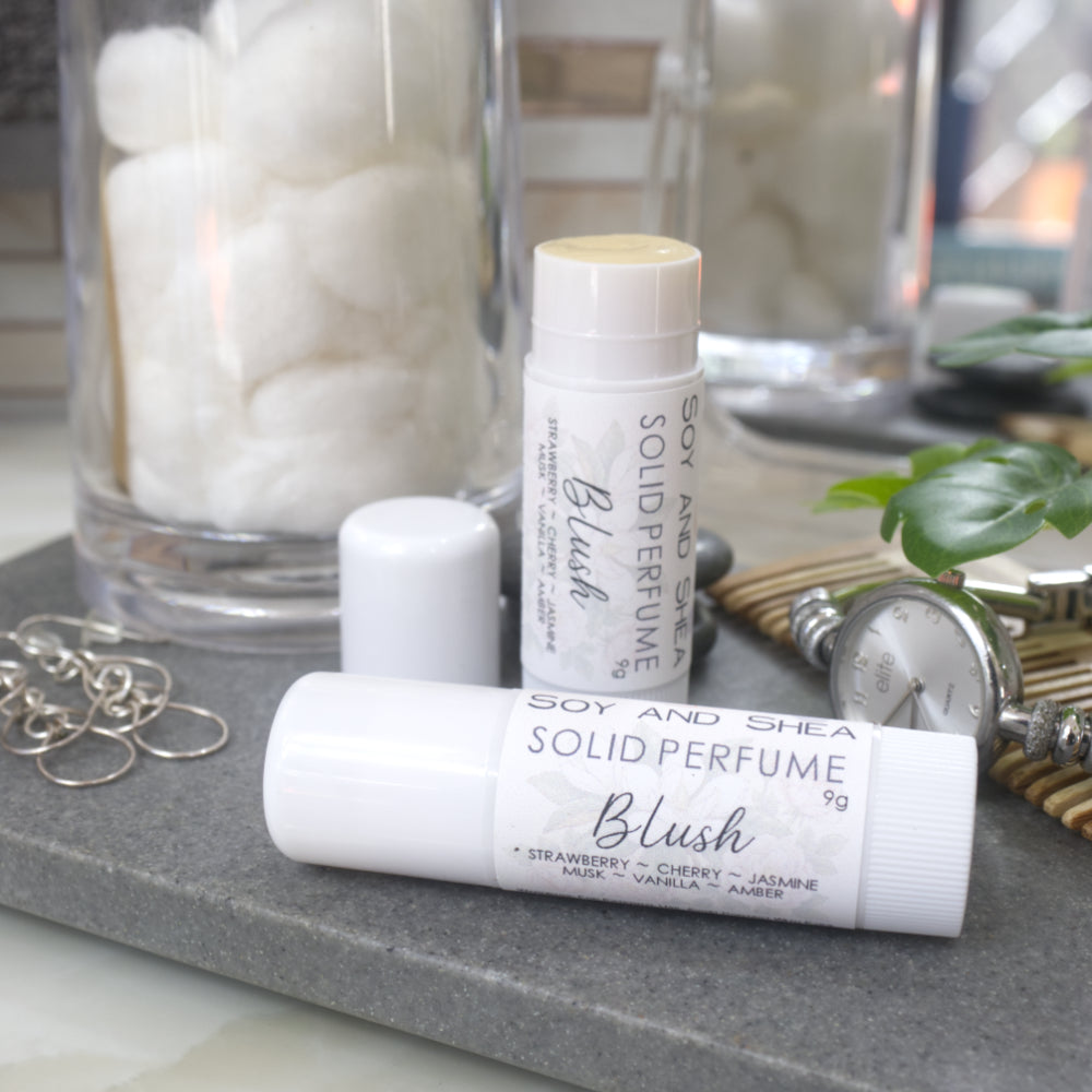 Two white tubes of Solid Perfume are displayed on a grey stone tray along with silver earrings, watch and greenery.   One tube lays flat  displaying the fragrance notes while another tube stands upright behind it with the lid removed and to the left revealing a beige balm