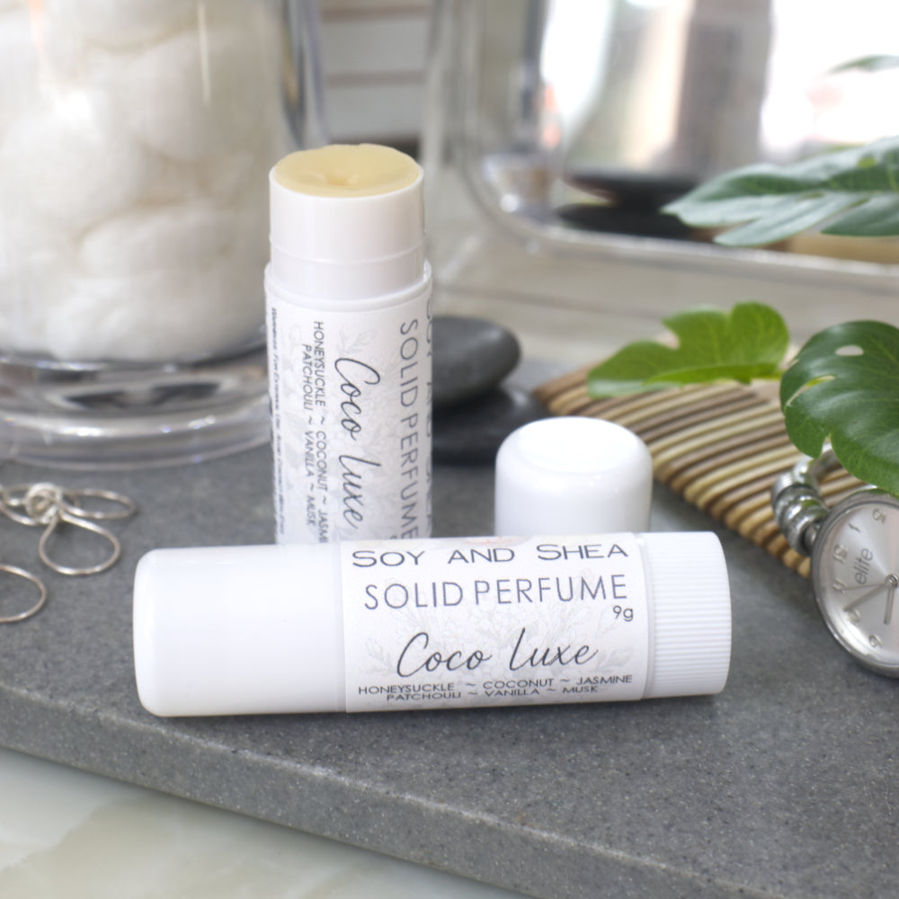 Two white tubes of Solid Perfume are displayed on a grey stone tray along with silver earrings, watch and greenery.   One tube lays flat  displaying the fragrance notes while another tube stands upright behind it with the lid removed and to the right revealing a beige balm