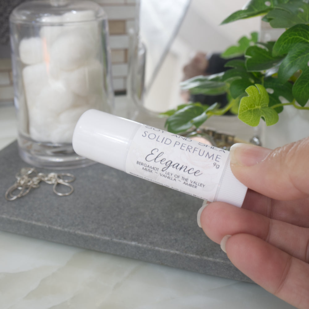 A hand holds a white wind up tube with a product label identifying it as Elegance Solid Perfume.  Behinds is a blurred bathroom countertop scene