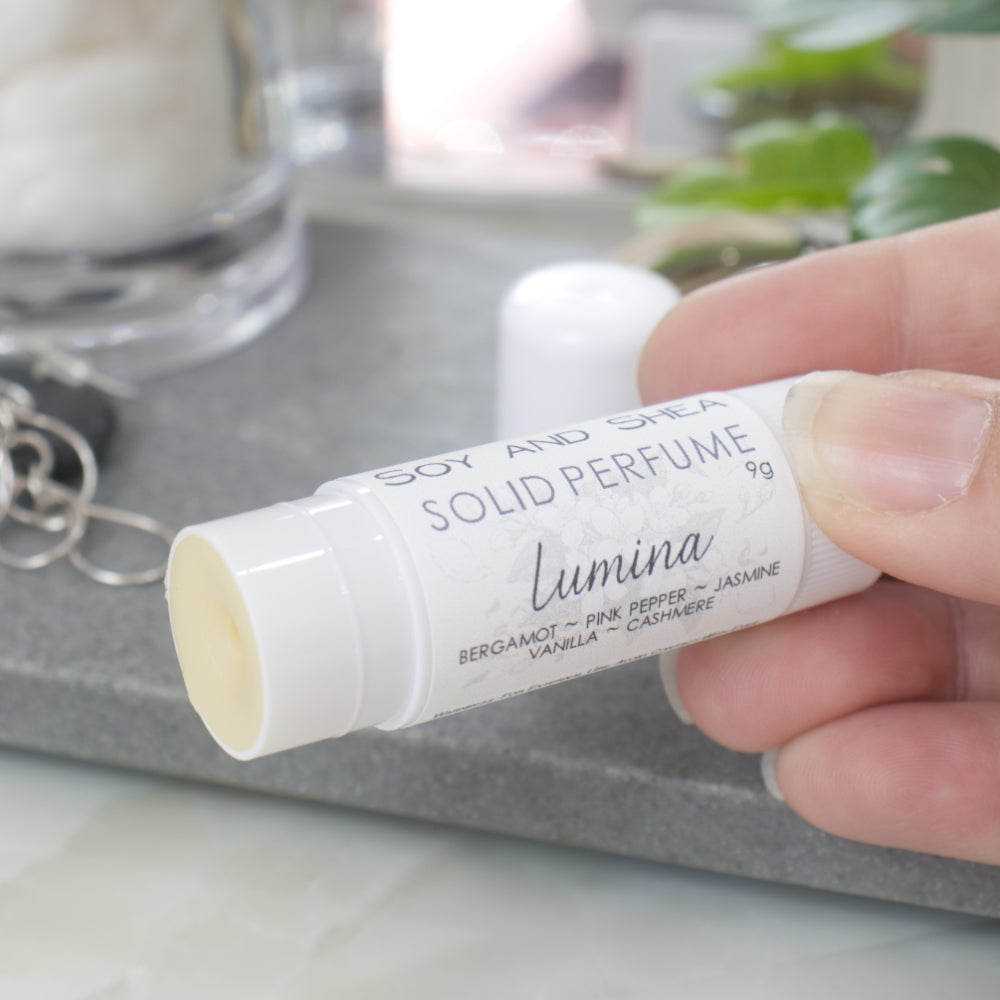 A hand holds a white wind-up tube labelled as Lumina Solid Perfume against a blurred background.  The lid has been removed to reveal a beige balm.