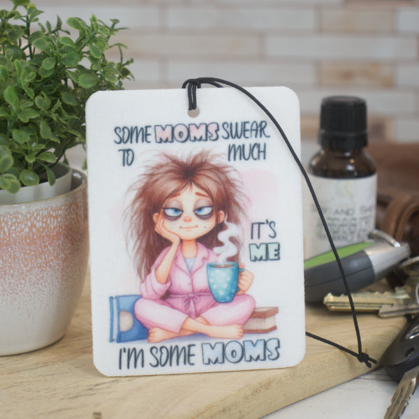 A car air freshener rests against a pot plant featuring and image of a cartoon mum sitting on the floor drinking coffee.  The air freshen says "Some Moms swear to much.  It's me.  I'm some moms"