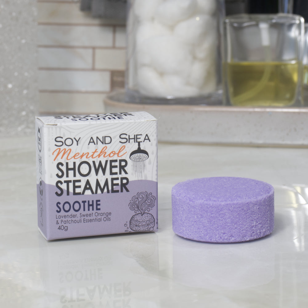 A boxed shower steamer and the steamer itself on a countertop in a bathroom setting. The box reads 'Soy and Shea Menthol Shower Steamer Soothe' and the round steamer is a purple color.