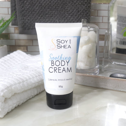 A white tube with black flip top cap stands on a bathroom counter next to a white towel and bathroom accessories.  The tube features a decorative product label with blue background announcing it as Soothing Body Cream with calendula, Arnica and menthol