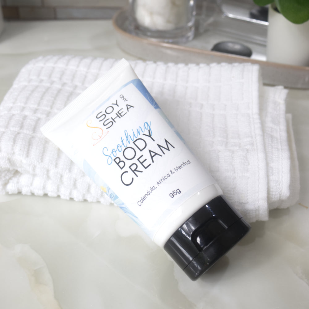 A white tube with black flip top cap lays on a bathroom counter against a white towel.  The tube features a decorative product label with blue background announcing it as Soothing Body Cream with calendula, Arnica and menthol