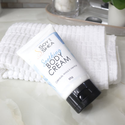 A white tube with black flip top cap lays on a bathroom counter against a white towel.  The tube features a decorative product label with blue background announcing it as Soothing Body Cream with calendula, Arnica and menthol