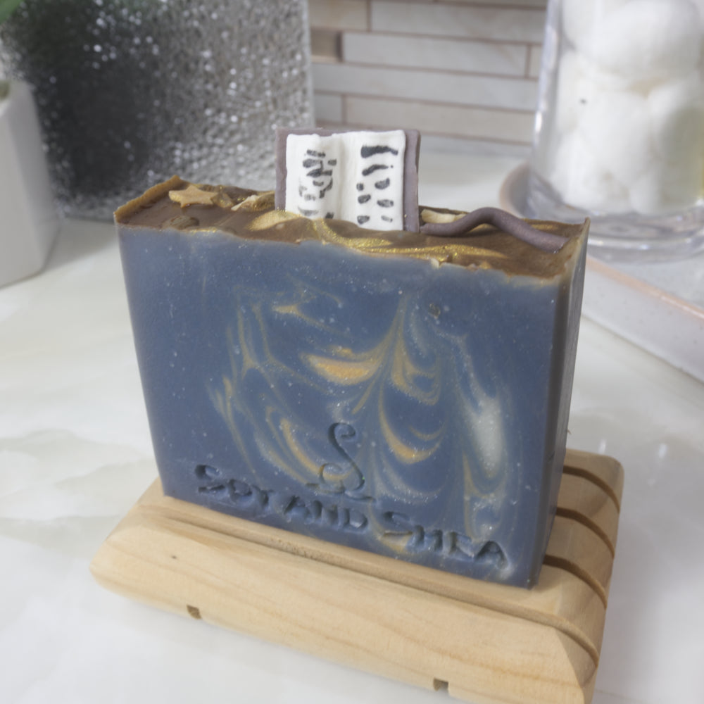 A soap bar stands on a wooden soap dish on a bathroom counter.  The soap is dark blue with gold and white streaks of colour throughout.  The top of the soap features embellishments made of soap including an open book standing up on the soap along with a wand and gold stars.