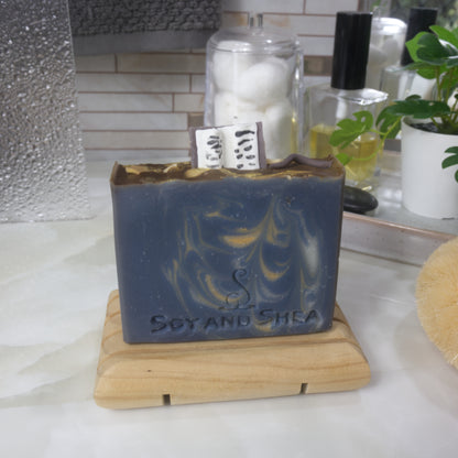 A soap bar stands on a wooden soap dish on a bathroom counter surrounded by perfume, jars and a plant.  The soap is dark blue with gold and white streaks of colour throughout.  The top of the soap features embellishments made of soap including an open book standing up on the soap along with a wand and gold stars.