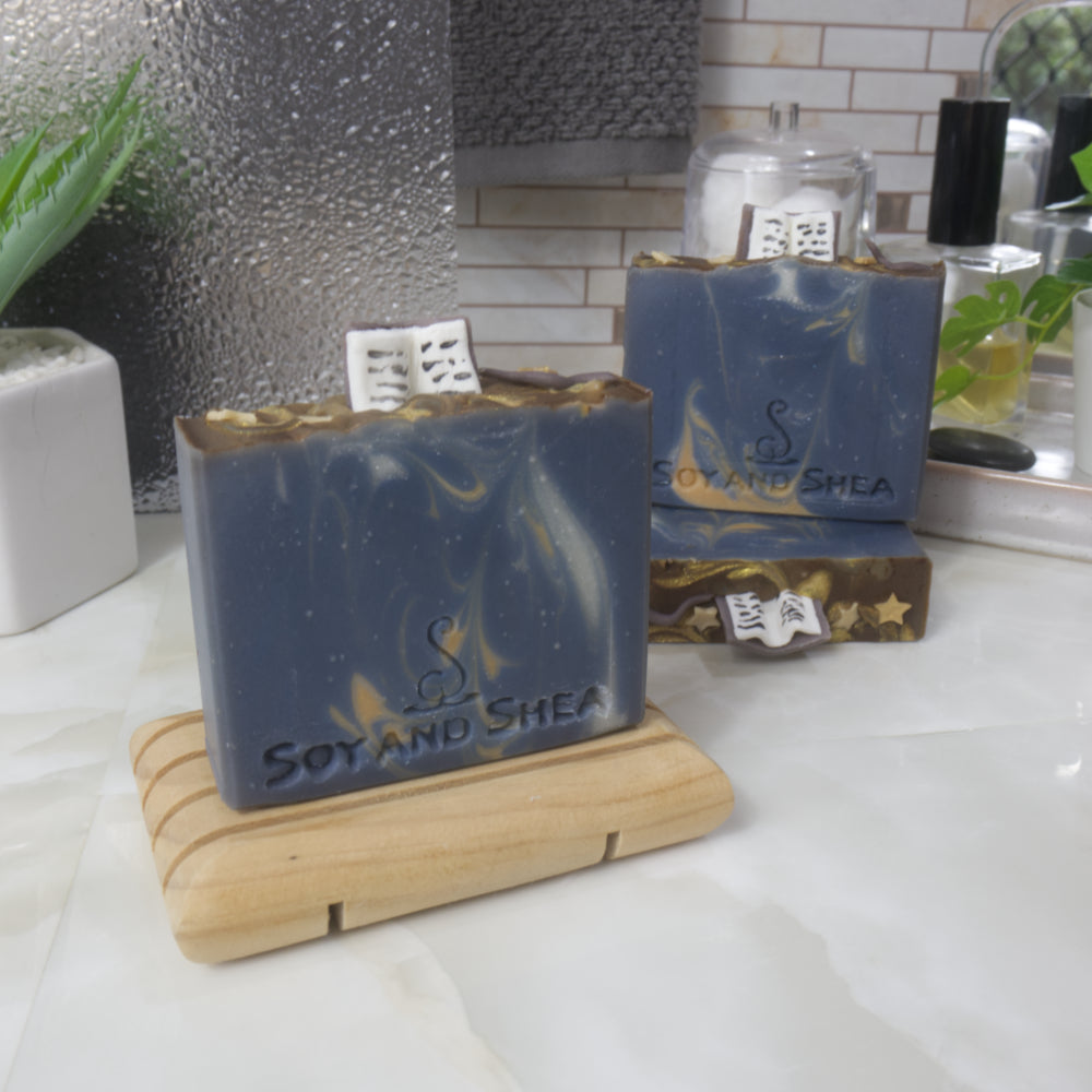 A selection of three soaps displayed on a bathroom bendch along with a bottle of perfume, plant and jar of cotton balls.  One soap sits on a wooden rack to the front left of the image, another two are displayed behind with one laying flat showing the top of the soap with a third standing on top.  The soaps are a dark blue with various swirls of gold and white and feature soap embellishment of an open book on top of each soap along with some stars and a wand.