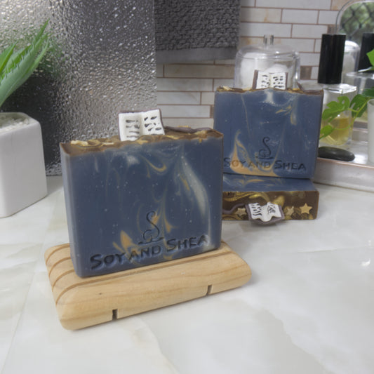 A selection of three soaps displayed on a bathroom bendch along with a bottle of perfume, plant and jar of cotton balls.  One soap sits on a wooden rack to the front left of the image, another two are displayed behind with one laying flat showing the top of the soap with a third standing on top.  The soaps are a dark blue with various swirls of gold and white and feature soap embellishment of an open book on top of each soap along with some stars and a wand.
