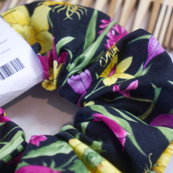 A detailed view of the Spring Bulbs Scrunchie by Soy and Shea, crafted from handmade cotton with a floral pattern showcasing pink, yellow, and green hues on dark fabric. Its knotless elastic design offers exceptional comfort. The packaging includes a visible barcode against a wooden background.