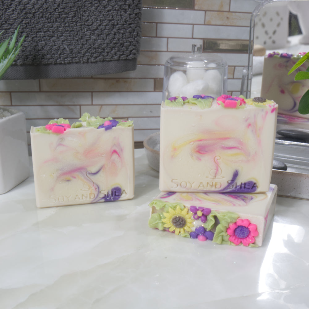 A collection of three soaps are displayed on a bathroom counter.  One soap stands upright to the back left, while to the front right, another stands on top of a soap lying down.  The flat soap shows a decorative top of brighty coloured flower embellishments.  All the soaps are a cream colour with varying swirls of neon pink, Bright yellow and dark purple.