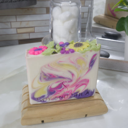 A rectangular bar of soaps stands on a wooden soap dish on a bathroom countertop.  The soap has a cream base colour with swirls in neon pink, dark purple and bright yellow.  The top features a collection of brightly coloured flower embellishments including a sunflower