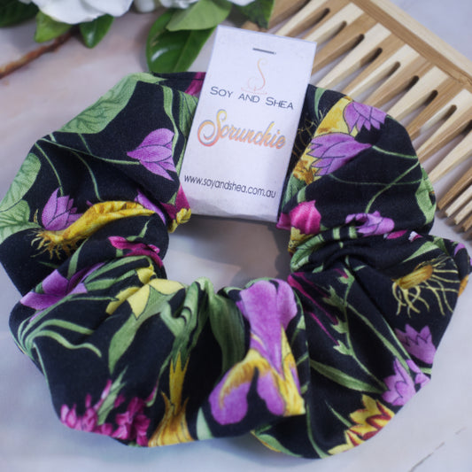 A colorful scrunchie sits on white surface resting against a wooden comb with greenery in the background.  A cardboard tag reads SOY AND SHEA SCRUNCHIE.    The fabric has a bold  print of purple and yellow spring bulb flowers with bright green leaves against a solid black background