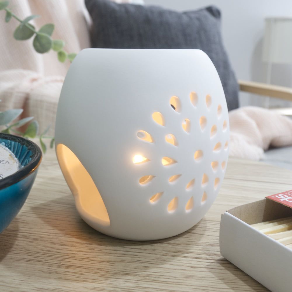 A smooth, matte white ceramic fragrance warmer sits on a light wooden table, glowing warmly from an internal tealight candle visible through a large, rounded opening. The side of the warmer features a circular starburst pattern of teardrop-shaped cutouts that let the candlelight shine through. To the left, a blue ceramic bowl holds wrapped wax melts, and a box of matches rests on the right. In the blurred background, a pink textured throw blanket and a dark grey cushion are draped over a chair.