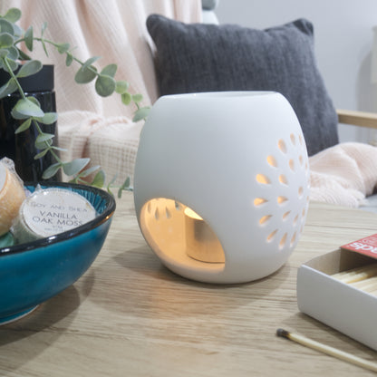A smooth, matte white ceramic fragrance warmer sits on a light wooden table, glowing warmly from an internal tealight candle visible through a large, rounded opening. The side of the warmer features a circular starburst pattern of teardrop-shaped cutouts that let the candlelight shine through. To the left, a blue ceramic bowl holds wrapped wax melts, and a box of matches rests on the right. In the blurred background, a pink textured throw blanket and a dark grey cushion are draped over a chair.