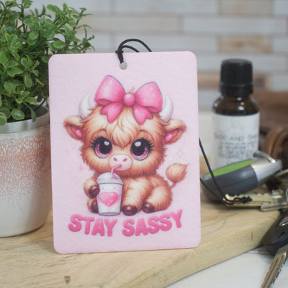 A car air freshener featuring an illustration of a cow with a pink bow and the phrase 'STAY SASSY' on a pink background.