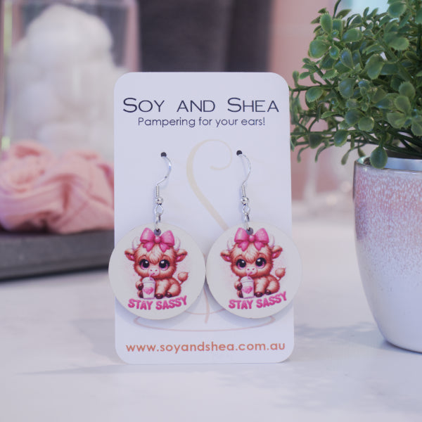 A pair of earrings on silver hooks hang on a white card branded with SOY AND SHEA leaning against a pot with a green leafy plant and a blurred background.  The earrings are round with a cartoon image of a baby highland cow with pink bow on head drinking from a cup with a heart and straw above the words STAY SASSY in bright pink