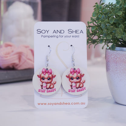 A pair of earrings on silver hooks hang on a white card branded with SOY AND SHEA leaning against a pot with a green leafy plant and a blurred background.  The earrings are round with a cartoon image of a baby highland cow with pink bow on head drinking from a cup with a heart and straw above the words STAY SASSY in bright pink