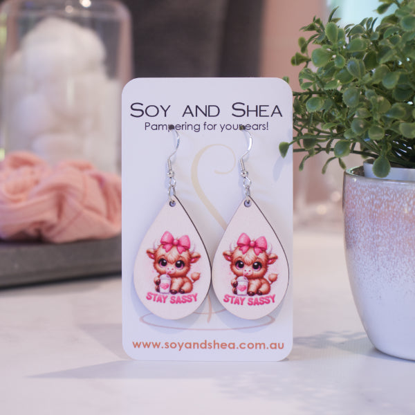 A pair of earrings on silver hooks hang on a white card branded with SOY AND SHEA leaning against a pot with a green leafy plant and a blurred background.  The earrings are teardrop shaped with a cartoon image of a baby highland cow with pink bow on head drinking from a cup with a heart and straw above the words STAY SASSY in bright pink