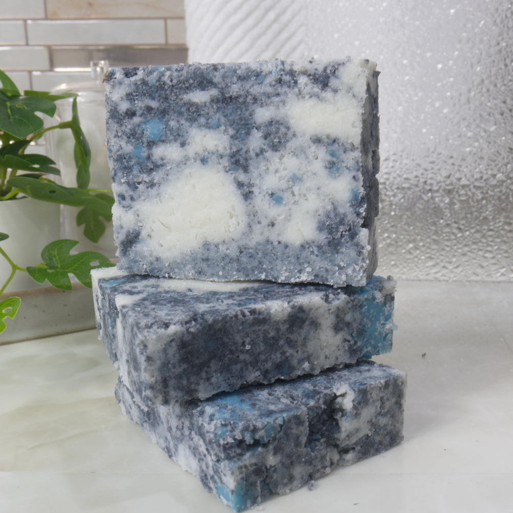 A selection of 3 soaps are stacked on a bathroom counter.  Two soaps lay flat with a third standing on top.  The soaps are a mottled design with grey, teal and white colours