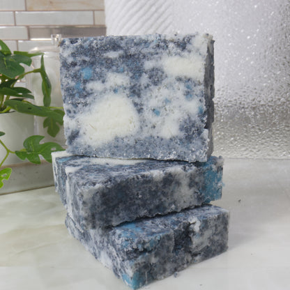 A selection of 3 soaps are stacked on a bathroom counter.  Two soaps lay flat with a third standing on top.  The soaps are a mottled design with grey, teal and white colours