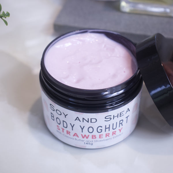 An open jar of Body Yoghurt sits on a marble top with a blurred background.  The lid rests on the side of the black jar revealing a pale pink cream