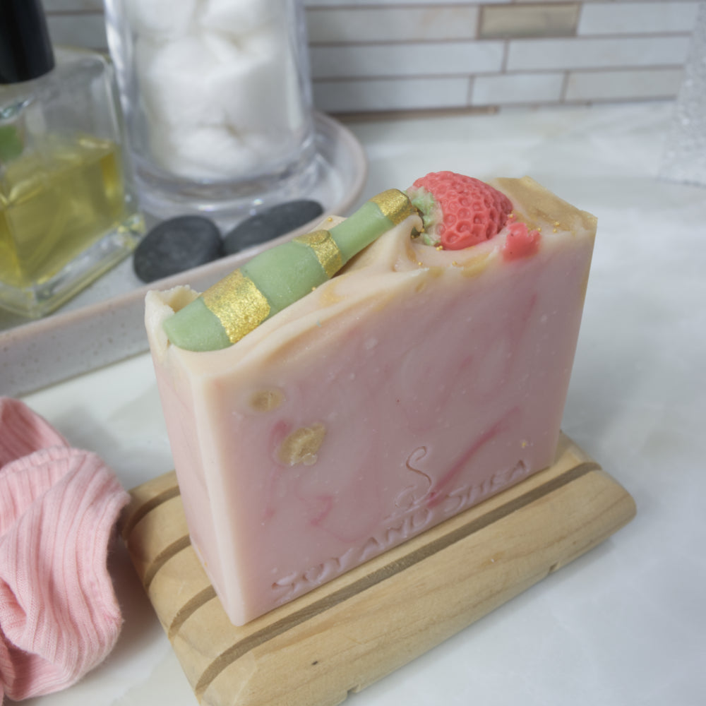 A bar of soap stands upon a wooden soap dish on a bathroom countertop.  The soap is pale pink with dark pink and gold swirls.  The top of the soap features soap embeds of a green champagne bottle and a red strawberry