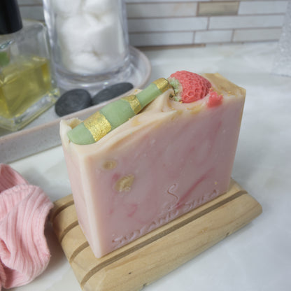 A bar of soap stands upon a wooden soap dish on a bathroom countertop.  The soap is pale pink with dark pink and gold swirls.  The top of the soap features soap embeds of a green champagne bottle and a red strawberry