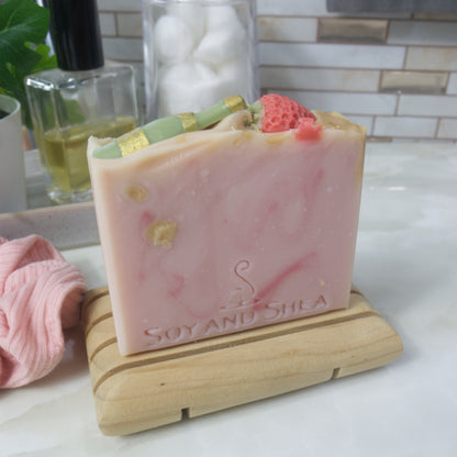 A bar of soap stands upon a wooden soap dish on a bathroom countertop.  The soap is pale pink with dark pink and gold swirls.  The top of the soap features soap embeds of a green champagne bottle and a red strawberry
